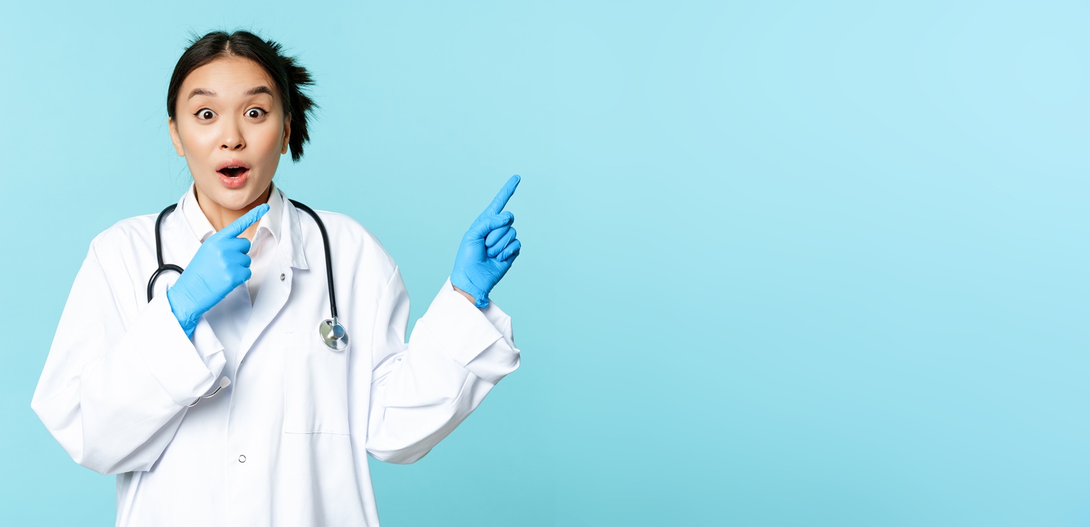 shocked asian doctor, nurse in uniform, pointing right and gasping surprised, big news, standing over blue background